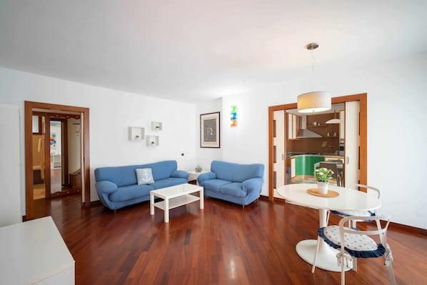 Ein Wohnzimmer mit blauem Sofa, einem weißen Couchtisch und einem Essbereich mit weißem Tisch und Stühlen.