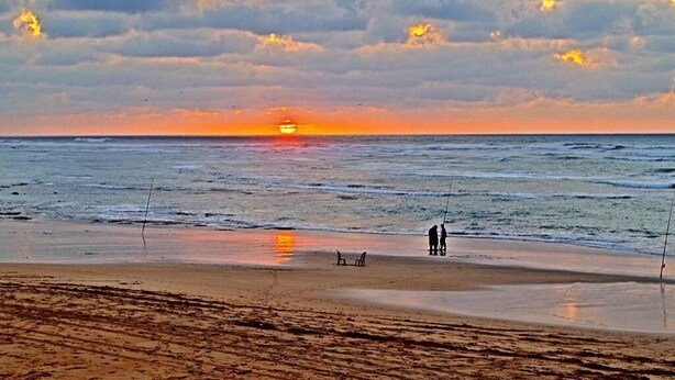 Une plage au coucher du soleil, deux personnes qui pêchent et une chaise sur le sable.