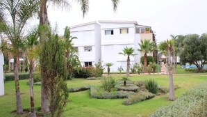 A modern building with a flat roof, surrounded by a well-maintained garden with various shrubs and palm trees.