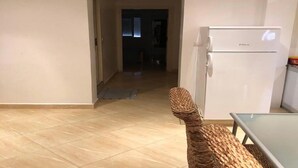 Ein Zimmer mit Fliesenboden, einem Korbstuhl, einem weißen Kühlschrank und einem Glastisch.
