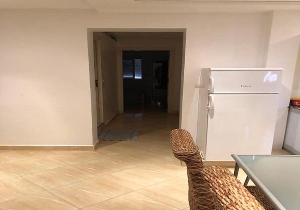 A room with a tiled floor, a wicker chair, a white refrigerator, and a glass table.