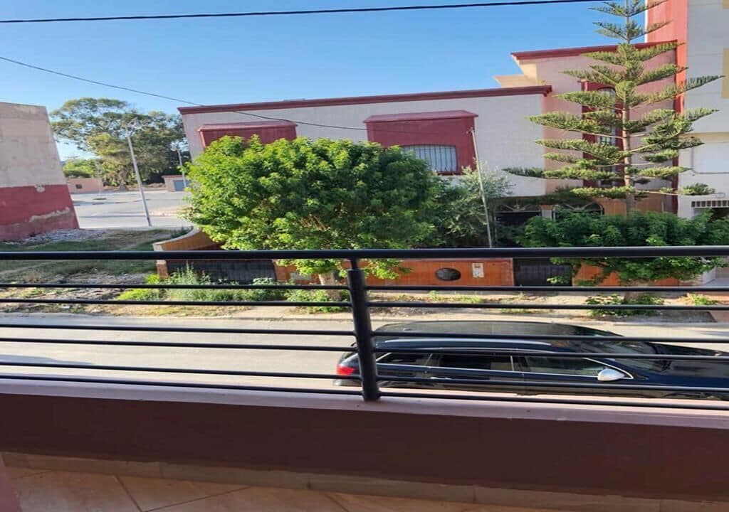 Un balcon donnant sur un quartier résidentiel, avec une voiture dans la rue et des immeubles aux façades rouges et blanches.