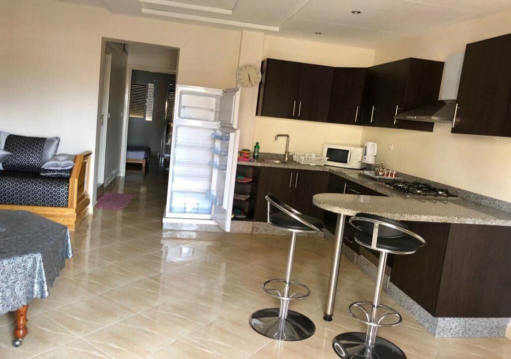 A modern kitchen with dark cabinets, a refrigerator, a microwave, and a gas stove. There are two bar stools and a small dining table with a tablecloth.