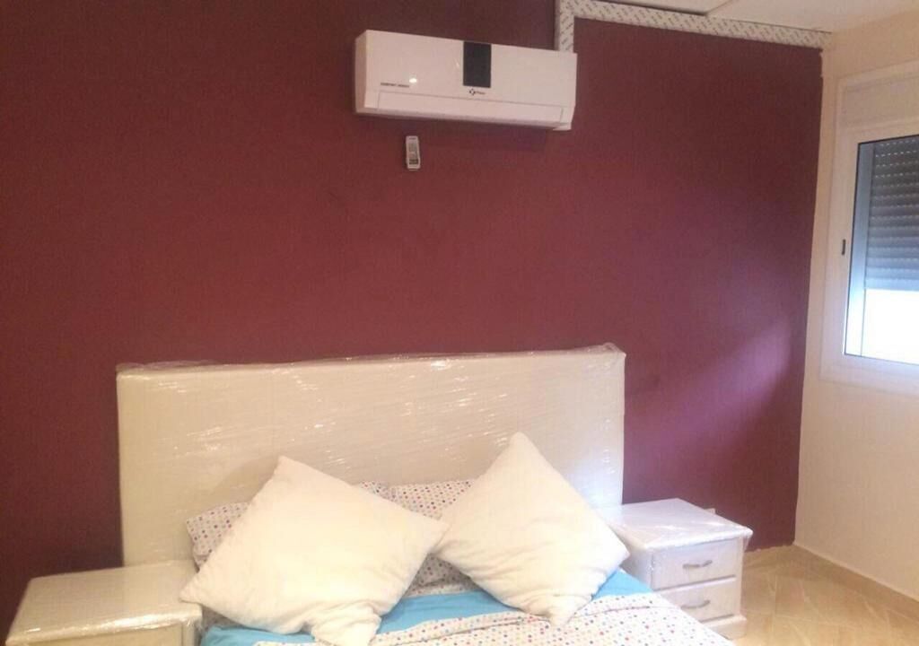 Une chambre à coucher avec un lit, une tête de lit blanche, deux oreillers, une table de chevet, une commode blanche et un climatiseur fixé au mur.