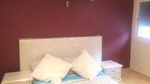 Een slaapkamer met een bed, een wit hoofdeinde, twee kussens, een nachtkastje, een witte dressoir en een wandairconditioner.
