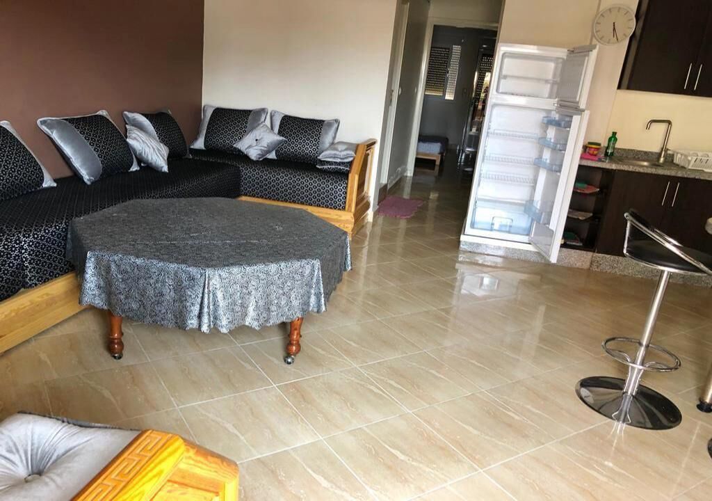 Ein Wohnzimmer mit Ecksofa, rundem Couchtisch, Barhocker und einer Küchenzeile inklusive Kühlschrank und Spüle.