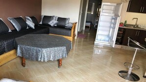 Een woonkamer met een hoekbank, een ronde salontafel, een barkruk en een keuken met een koelkast en gootsteen.