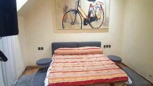A bedroom with a bed, a small stool, a wall-mounted bicycle painting, and a window.