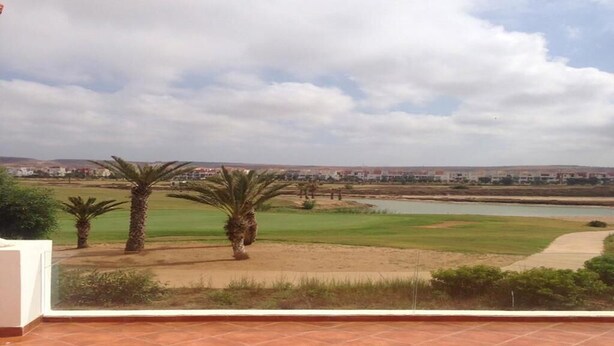 A view from a balcony with a tiled floor, overlooking a golf course with palm trees and a body of water. - Superb Villa in Marina Saidia 4899 (Saidia)