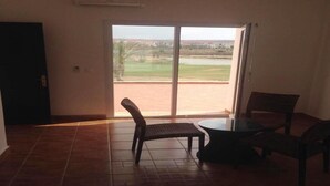 A room with a tiled floor, a sliding glass door leading to a view of a golf course, a small round table, and two chairs. - Superb Villa in Marina Saidia 4899 (Saidia)