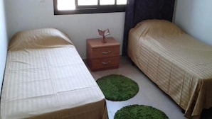 Une petite chambre propre et bien rangée, avec deux lits simples, une table de chevet en bois et une fenêtre avec des rideaux.
