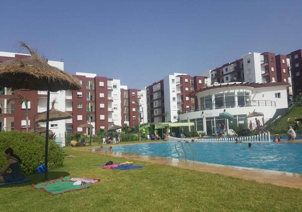 A swimming pool area with a thatched umbrella, a grassy lawn, and multi-story buildings in the background.