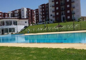 A swimming pool with a grassy area in front, surrounded by multi-story residential buildings.