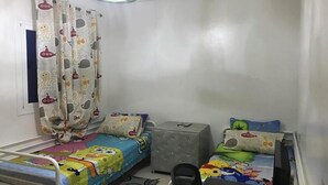 Een kleine, goed verlichte kamer met twee bedden, waarvan één een kleurrijke deken met tekenfilms en de andere een effen witte deken. Er staat een klein grijs kastje en een zwarte stoel in de kamer.