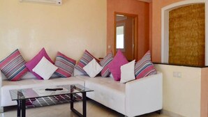 A living room with a white sofa, striped and solid-colored cushions, a glass coffee table, and an air conditioning unit on the wall. - Pleasant sea view apartment El Hamra II 4919 (Asilah)