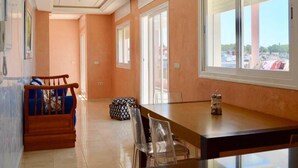 A hotel room with a wooden dining table, clear chairs, a patterned ottoman, and large windows offering a view of the parking lot. - Pleasant sea view apartment El Hamra II 4919 (Asilah)