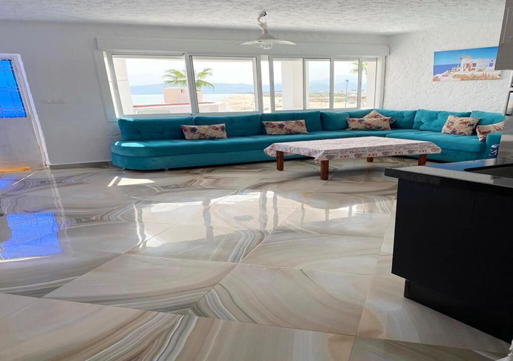 A living room with a teal sofa, a coffee table, and a large window with a view of the beach.