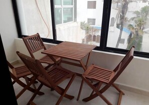 A balcony with a wooden table and chairs, large windows, and a view of buildings and palm trees. - Al Amine Beach Complex Fnideq 4928 (Fnideq)