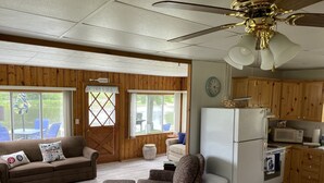 A cozy cabin interior with a living room, dining area, and kitchen, featuring wooden walls, a ceiling fan, and a view of the outdoors.