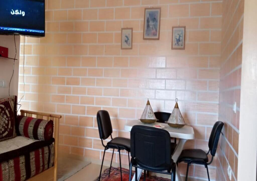 A room with a brick wall, a TV mounted on the wall, a sofa, a dining table with chairs, and framed pictures on the wall.