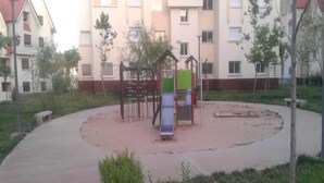 A playground with a slide and climbing structures, surrounded by a paved area and trees, with apartment buildings in the background. - Beautiful apartment in Les Jardins d'Ifrane 4930 (Ifrane)