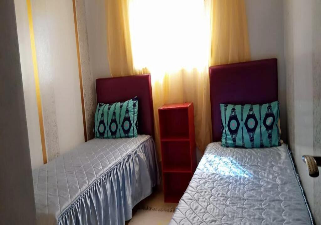 A room with two beds, each with patterned pillows, a red nightstand, and a window with curtains.
