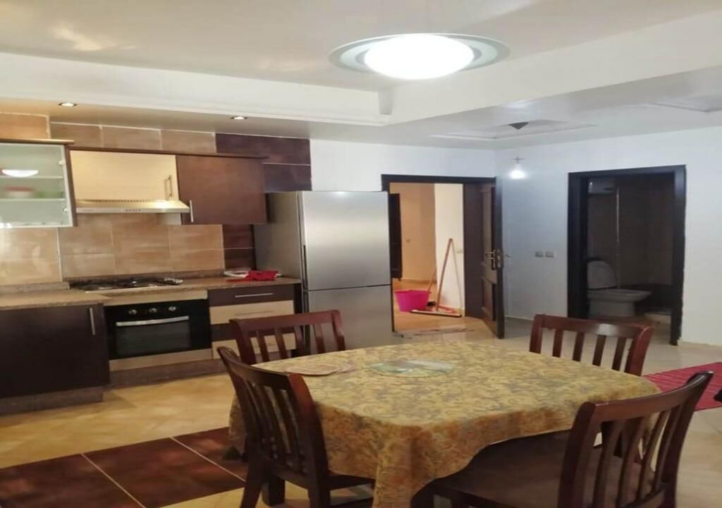 A dining area with a table and chairs, a kitchen with a stove and refrigerator, and a door leading to another room.