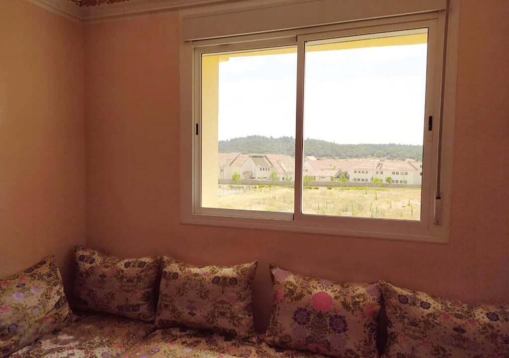 Una habitación con un ventanal grande, un sofá con cojines estampados y vistas a edificios y vegetación.
