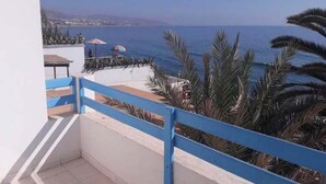 Un balcon doté d’une balustrade bleue, donnant sur une plage bordée de palmiers et surplombant un ciel dégagé.