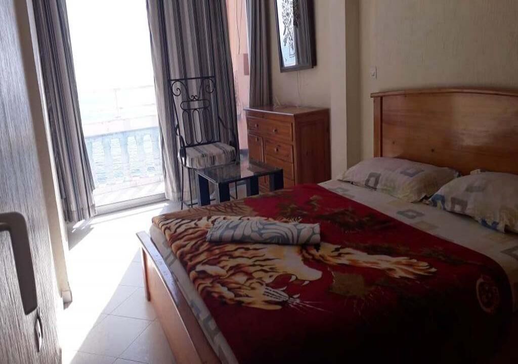Un dormitorio con cama, cabecera de madera, mesita de noche, una silla y una ventana con cortinas.