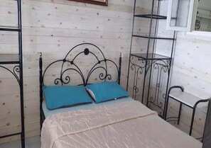 Uma cama de solteiro com estrutura metálica e colchão bege, duas almofadas azuis e uma parede revestida de madeira com uma pequena prateleira acima.