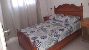 A single bed with a floral bedspread, a wooden headboard, a nightstand, and a window with curtains. - Pleasant Sea Front Apartment in Taghazout 4966 (Taghazout)