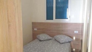 Une chambre à coucher avec une tête de lit en bois, deux oreillers, une couvre-lit à motifs, une fenêtre avec des stores et une petite table de chevet.