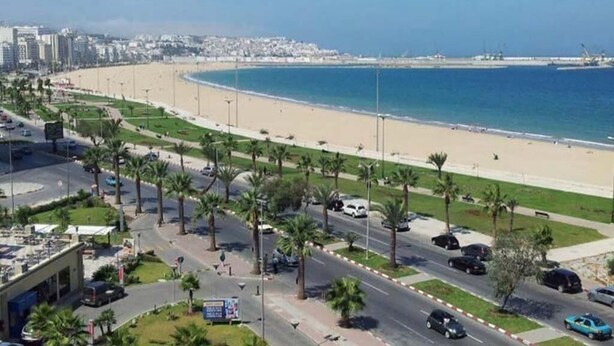 A coastal road with palm trees, a beach, and a cityscape in the background. - Superb apartment in Le Printemps 4972 (Tanger)