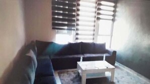 A living room with a dark-colored sofa, a white coffee table, and a window with blinds. - Superb apartment in Le Printemps 4972 (Tanger)