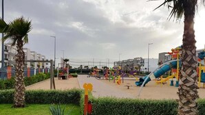 A playground with various play structures, including slides and climbing areas, surrounded by palm trees and a well-maintained lawn.