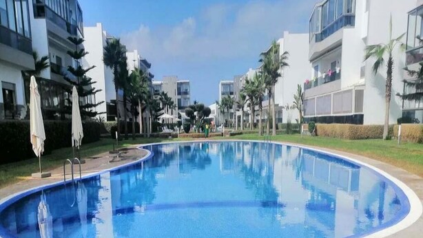 A swimming pool surrounded by modern buildings, palm trees, and a clear sky.