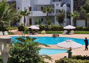 Un espace piscine doté de chaises longues et de parasols, entouré de palmiers et bordé d’un bâtiment avec des balcons.