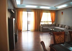 A spacious living room with a sofa set, a dining table, and a kitchen area visible in the background.