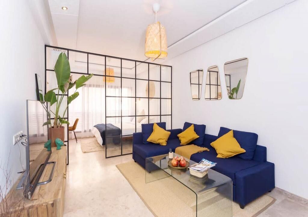 Una sala moderna con un sofá azul, cojines amarillos, una mesa de centro de vidrio y una planta grande.