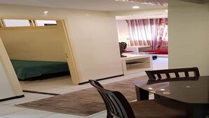 Een kamer met een eettafel en stoelen, een bed met een groene spreidekking, en een zithoek met een bank en gordijnen.