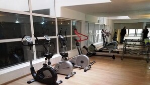 Academia com diversos equipamentos de exercício, amplas janelas e piso de madeira.