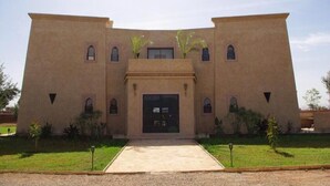 A large, beige building with a central entrance, multiple windows, and a small garden area with plants. - Pleasant villa with swimming pool in Marrakech 5100 (Marrakech)