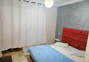 Quarto com cama, cortinas brancas, cabeceira vermelha e parede cinza.