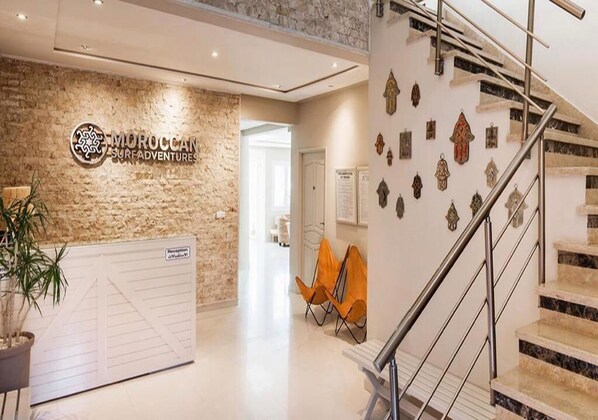 A reception area with a Moroccan Surf Adventures sign, a reception desk, a staircase, and a wall with decorative items.