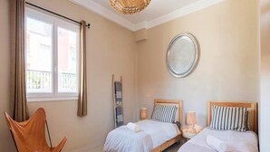 Ein Schlafzimmer mit zwei Betten, einem Sessel, einem runden Spiegel und einem Fenster mit Vorhängen.