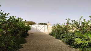 Un camino de grava que lleva a una puerta blanca, con un muro de piedra y una pequeña estructura parecida a una torre.