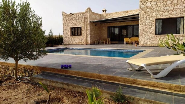 Una casa de piedra moderna con piscina, mobiliario de exterior y un pequeño árbol.