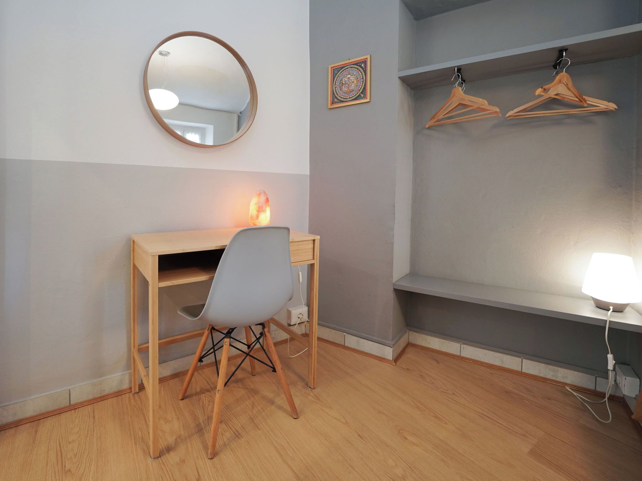 Een kamer met een houten bureau, een stoel, een ronde spiegel, een verwarming, een wandplank met houten kapstokken, en een bedlampje.