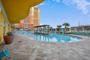 Zona de piscina con área de descanso techada, sillones reclinables y un edificio alto al fondo.